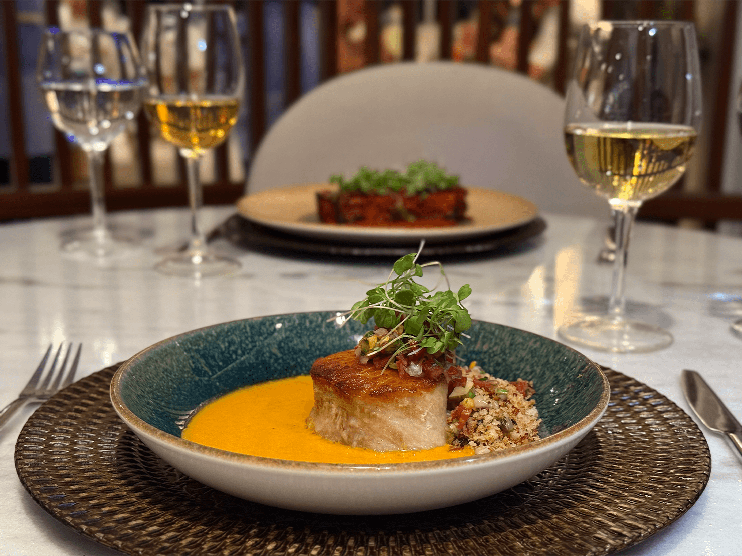 Prato com peixe grelhado, creme laranja e farofa crocante em mesa posta no Japaratinga Lounge Resort. Ao fundo, taças de vinho branco e ambiente aconchegante.