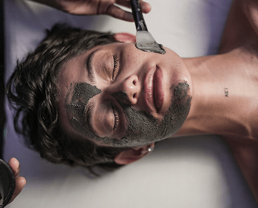 Jovem com uma máscara de argila no rosto sendo aplicada com um pincel, está recebendo um ritual facial.