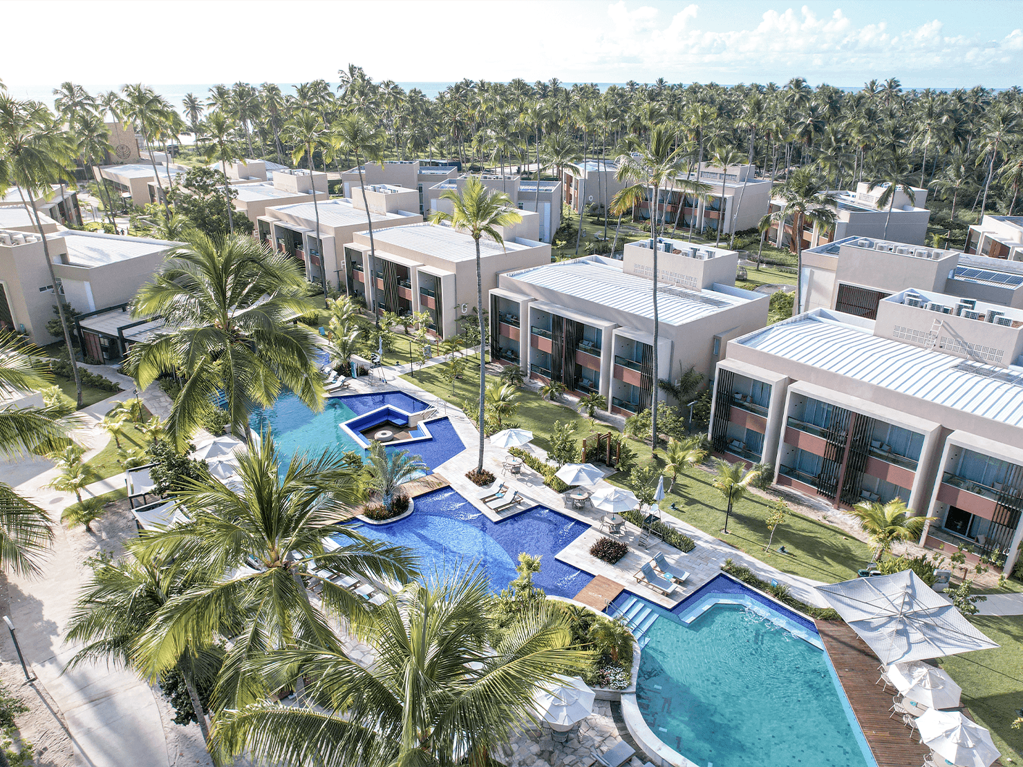 Vista aérea do Japaratinga Lounge Resort cercado por coqueiros, com piscinas em tons de azul, espreguiçadeiras e Gardens.