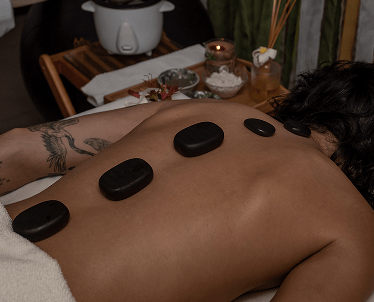 Mulher de costa em cama de massagem, com pedras pretas nas costas, ao redor tem utensílios de massagem.