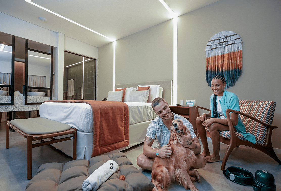 Casal na Garden Exclusive com cachorro no quarto, o homem está sentado no chão alisando o dog, e a mulher sentada na poltrona olhando pra eles e sorrindo, o quarto é moderno com decoração colorida.