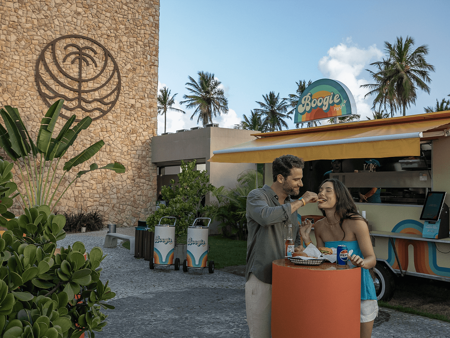 Casal jovem sorrindo e compartilhando um lanche em frente ao Boogie Truck, o food truck retrô do Japaratinga Lounge Resort. Ambiente externo com palmeiras, design vintage