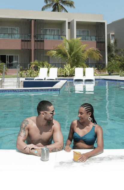 Casal na piscina na área das Gardens, com estrutura dos quartos ao fundo. Tem espreguiçadeiras e guarda-sóis ao redor da piscina e coqueiros altos.