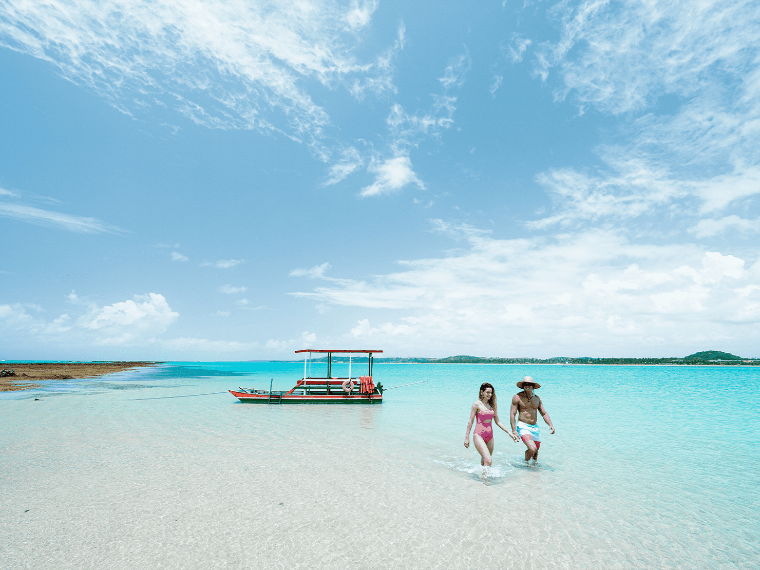 Casal caminhando em águas cristalinas e rasas na Croa de São Bento, próximo ao Japaratinga Lounge Resort. Barco rústico ancorado em banco de areia sob céu azul com nuvens brancas.